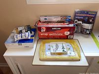 Wide view showing plastic wrap container (partial), aluminum foil box, placemat, plastic trays on white table against beige wall, and boxed beverage holder