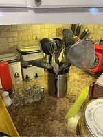 Kitchen countertop showing a stainless steel utensil holder filled with various kitchen utensils including spatula, whisk, ladle, tongs, and skimmer, beside multiple glass salt and pepper shakers and two kitchen scales.