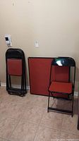 Four black metal folding chairs with red vinyl seat and back, folded flat and resting against a wall next to a card table.