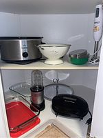All items displayed inside white cabinet including Rival Crock-Pot, Magic Bullet blender, Cuisinart hand blender, glass baking dishes, strainer, cheese grater, serving tray, and Hamilton Beach mini grill.