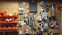 Wide shot showing multiple orange bins with screws, grommets, and metal hardware hung on pegboard showing saws, callipers, tapes, clipboard, and bird feeder part