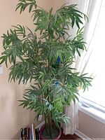 Full view of tall artificial bamboo plant with green leaves and brownish-green stems in a red round pot near window and books.