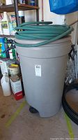 Overall view of gray Rubbermaid garbage can with green hose coiled on top in a basement.