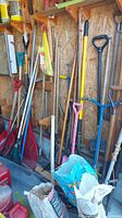 Wide view of various garden tools hung against a plywood wall including rakes, shovels, broom, and bags of materials stored at the base.
