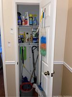 Full view of closet with hanging and shelf-stored cleaning supplies, bucket, mops and broom inside a small closet