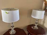 Pair of crystal table lamps with white fabric drum shades and silver bases on wooden surfaces.