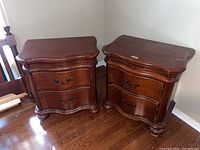 Pair of matching wooden night tables with two drawers and pull-out shelves, shown side by side in corner near a bed frame on hardwood floor.