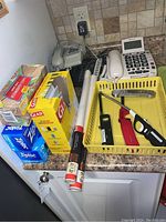 Two Bell telephones, one ClearSounds telephone with large buttons, partial boxes of Ziploc and Glad bags, two packs of shelf liner, three BBQ lighters, a meat thermometer, and yellow plastic tray on kitchen counter.