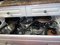 Drawer containing multiple pots and frying pans, some with lids stored inside. Variety of sizes and materials.