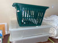 Photo of a dark green plastic laundry basket placed on top of two clear plastic storage bins with lids.