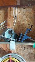 Wide view of a wooden shelf with two metal vices positioned side-by-side, showing a larger bench vise and smaller clamp-on vice.