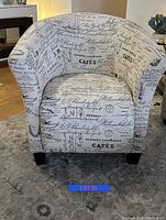 Front view of a barrel-style accent chair upholstered in white fabric with printed black script and French café-themed text, standing on a carpeted floor.