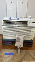 Front view of LG window air conditioner showing air vents and attached power cord. Owner's manual and remote controller placed in front.