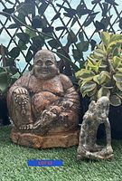 Front view showing Buddha statue and smaller stone seated figure statue positioned on grass with plants in background