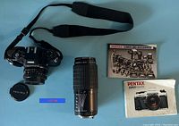 Pentax Program Plus camera body with attached 50mm lens, Takumar-A 70-200mm zoom lens, Pentax lens cap, camera strap, and two manuals on light blue surface