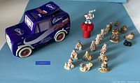 Dark purple Cadbury tin truck with red and white wheels, Cadbury logo and Frigidaire text visible. Next to it, red Hershey's sticker dispenser figurine holding a bunny on top. Multiple Red Rose Tea figurines in cream and brown shades are aligned, some with visible chips.