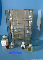 Photo of five miniature fragrance bottles in front of empty glass and brass curio cabinet with a front door open against blue background.