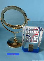 Front view of brass tabletop swivel mirror and acrylic Eaton's 125 Year Anniversary paperweight with a miniature house inside, displayed on a blue background.