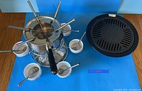 Photo showing the 6-person fondue set with forks, ceramic bowls and the separate roasting tray on a wooden surface against a blue background.