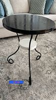 Round metal side table with black perforated top, curled black metal legs, white lower shelf on carpeted floor