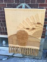 Full view of the beige and brown wool wall hanging with fringe, showing textured abstract shapes representing an oval form and fan-like rays.