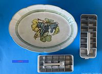 Top view of the large ceramic serving platter with colorful grape design in center, accompanied by two vintage metal ice cube makers with handles on a blue background.