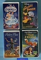 Four VHS tapes with Disney-themed illustrated cardboard sleeves on a blue surface, showing the front covers for Cinderella, The Brave Little Toaster, Peter Pan, and The Fox and the Hound.
