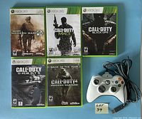 Front view of five Xbox 360 Call of Duty games cases and white wired Xbox 360 controller arranged on flat surface showing cover art.