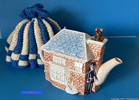 Side view of the porcelain house-shaped teapot with a female mouse figurine in a white dress, a male mouse figurine in black attire by the door, and a mouse atop the chimney. Blue tiled roof and detailed brickwork visible.