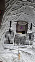 Four BBQ grill baskets laid out on white fabric background, three wire mesh rectangular baskets and one perforated metal square basket with various handles.