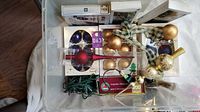 Photo showing multiple boxed sets of Christmas ornaments including gold balls, purple cones, and red finials, plus a string of green wire Christmas lights and beaded garland with ribbon inside a plastic bin.