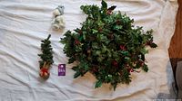 Photo showing a small artificial Christmas tree, a snowman figurine, and a holly wreath laid out on a white cloth background.