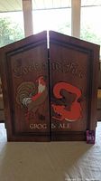 Closed wooden dartboard cabinet with painted rooster and fox and text 'Cock and Fox Grog & Ale'