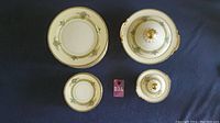 Overview of two dinner plates and two lidded serving vessels with floral and gold accents on a dark blue cloth.