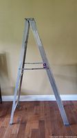 Side-angle view of six-foot aluminum ladder standing on wooden floor against a wall.