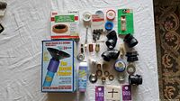 Various plumbing tools and supplies including One Second Plumber kit, toilet wax ring, tapes, black plastic fittings, brass plumbing fittings and adapters on white cloth background.