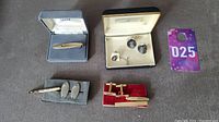 Four tie clips and three sets of cuff links arranged on a dark carpet surface, some in boxes, showing the full contents of the lot including the monogrammed cuff links.