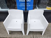Front view of two white plastic armchairs showing armrests, seat and backrest, placed side by side on a sidewalk.