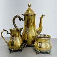 Three-piece vintage brass tea and coffee set with gold polish and engraved decorative bands, standing on ornate silver-tone feet.