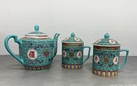 Front view of the teal and gold decorated teapot with two lidded cups, showing the detailed patterns and Chinese characters on all items.