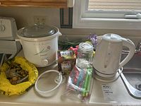 Photo of white Sunbeam electric kettle with water level indicators and corded base on a kitchen counter next to a Proctor Silex slow cooker and miscellaneous items.