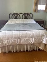 Front view of the bed showing the wooden carved headboard, metal frame, white mattress topper, white bed skirt, and gray throw blanket with one wooden nightstand on each side.