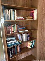 Four shelves filled with a diverse collection of books including vintage and modern titles shown from front view on wooden shelving unit.