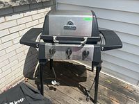 Front view of stainless steel Broil King gas barbecue with closed lid, black side shelves, and three control knobs. Black grill cover visible beside the barbecue.