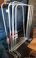 Six white metal standing clothing racks grouped together, positioned in a garage with blue tarp in background.