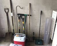 All garden tools including spades, rakes, shovels, shepherd's hook, sweeper, cooler, and rolled plastic mat arranged in garage against wall.
