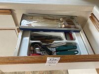 Drawer containing assorted kitchen utensils such as whisks, can opener, garlic press, and various spoons and spatulas in a white organizer.