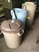 Three Rubbermaid garbage bins with lids in beige/light colors, one with black handles and wheels, one with brown lid, placed in a garage space