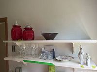 Shelf with two red glass canisters with lids, a dark pottery bowl, a decorative plate, six stem wine glasses, and two smaller clear glasses