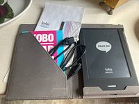 Photo showing Kobo e-reader in gray protective cover along with USB charging cable and Kobo manuals/booklets.